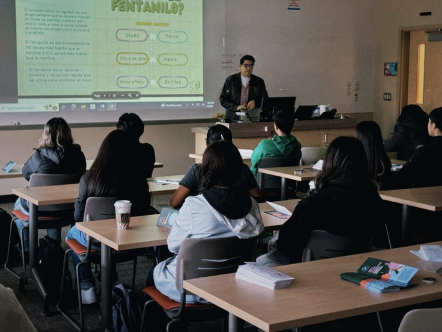 CHWCMR_2 A photo of students in a classroom listening to a speaker. A projector screen displays a Spanish-language presentation. Pamphlets are visible on the desks.
