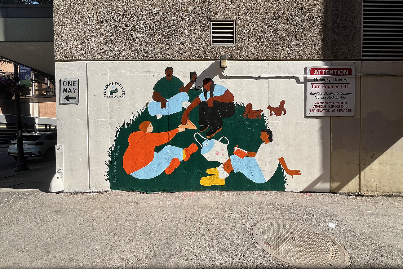 Colorful public mural of a diverse group sitting together in grass around tote bag with naloxone, with squirrels nearby, painted on a white city wall. 