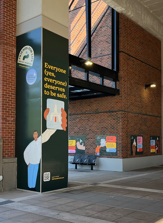 Brick transit station with a column wrapped in a Friends for Life naloxone campaign and additional colorful safety posters above a row of benches. 