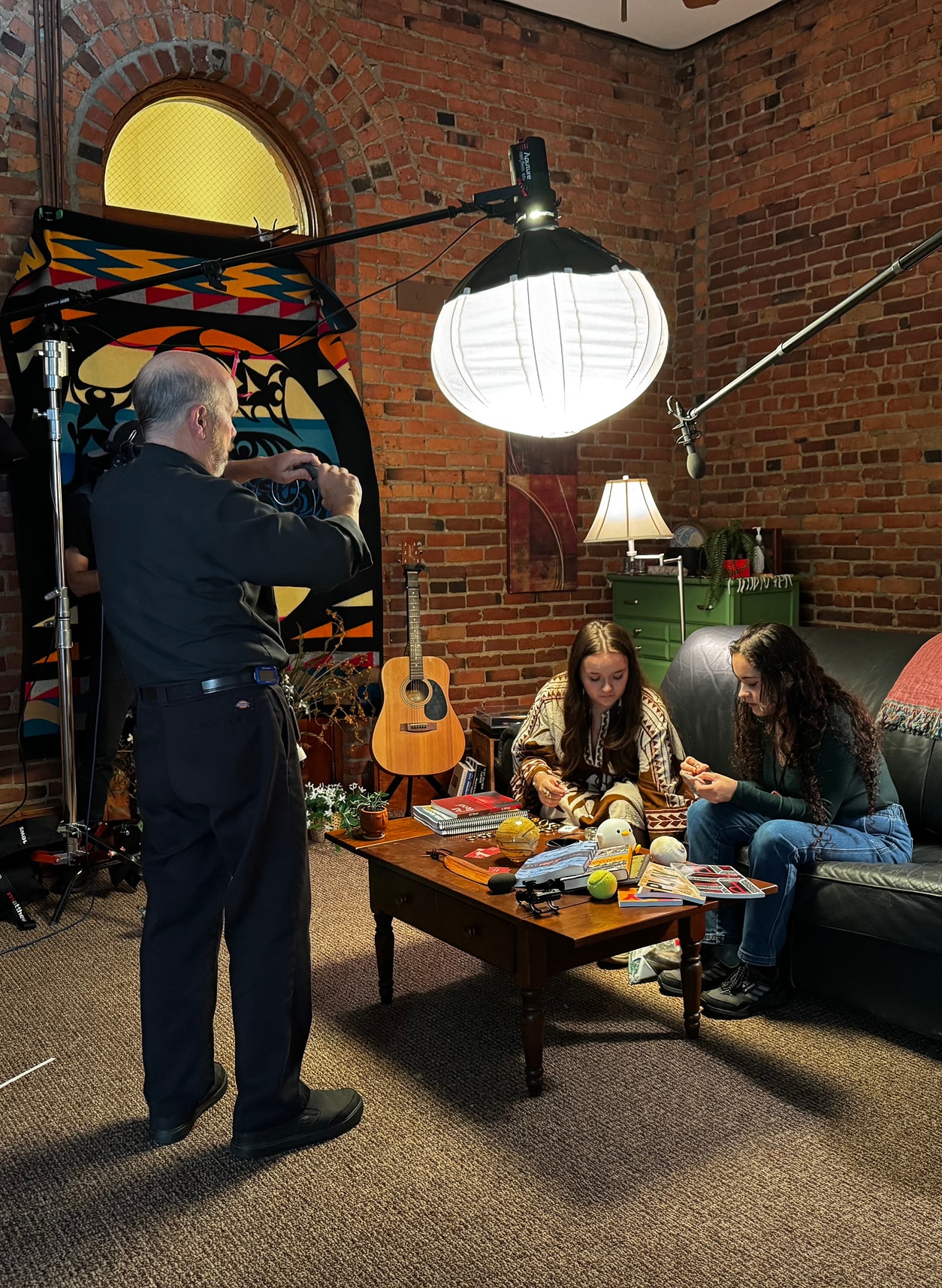 A behind-the-scenes photo from a video shoot in a brick-walled room. A crew member adjusts camera gear under a large studio light while Ayasha and Meera Forespring sit on a couch beading together. 