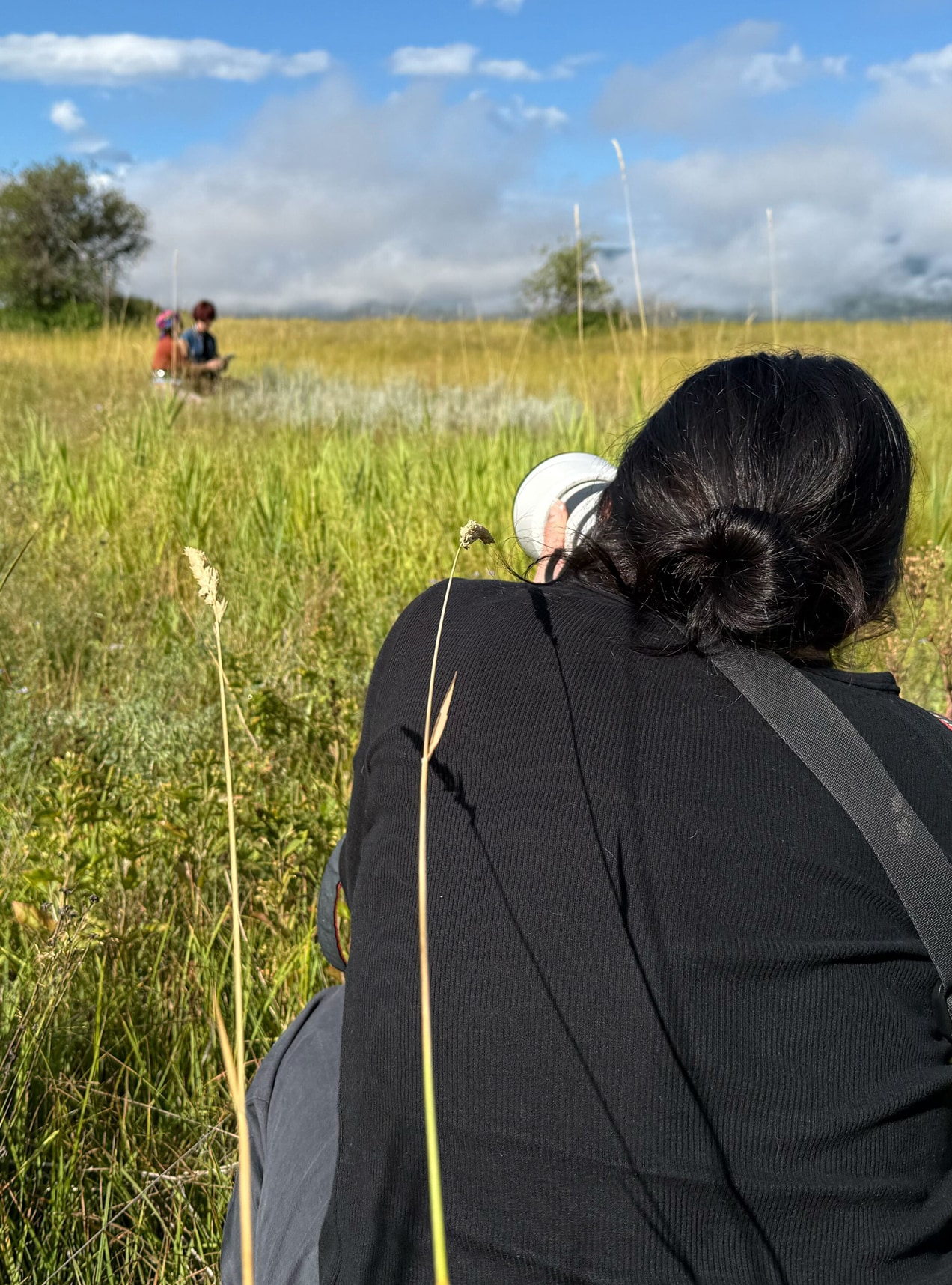 A behind-the-scenes photo from a photo shoot in a grassy meadow. Photographer Tailyr Irvine crouches in the tall grass, photographing Neomia Battin and his auntie Alisha Athos as they sit together in the distance. 