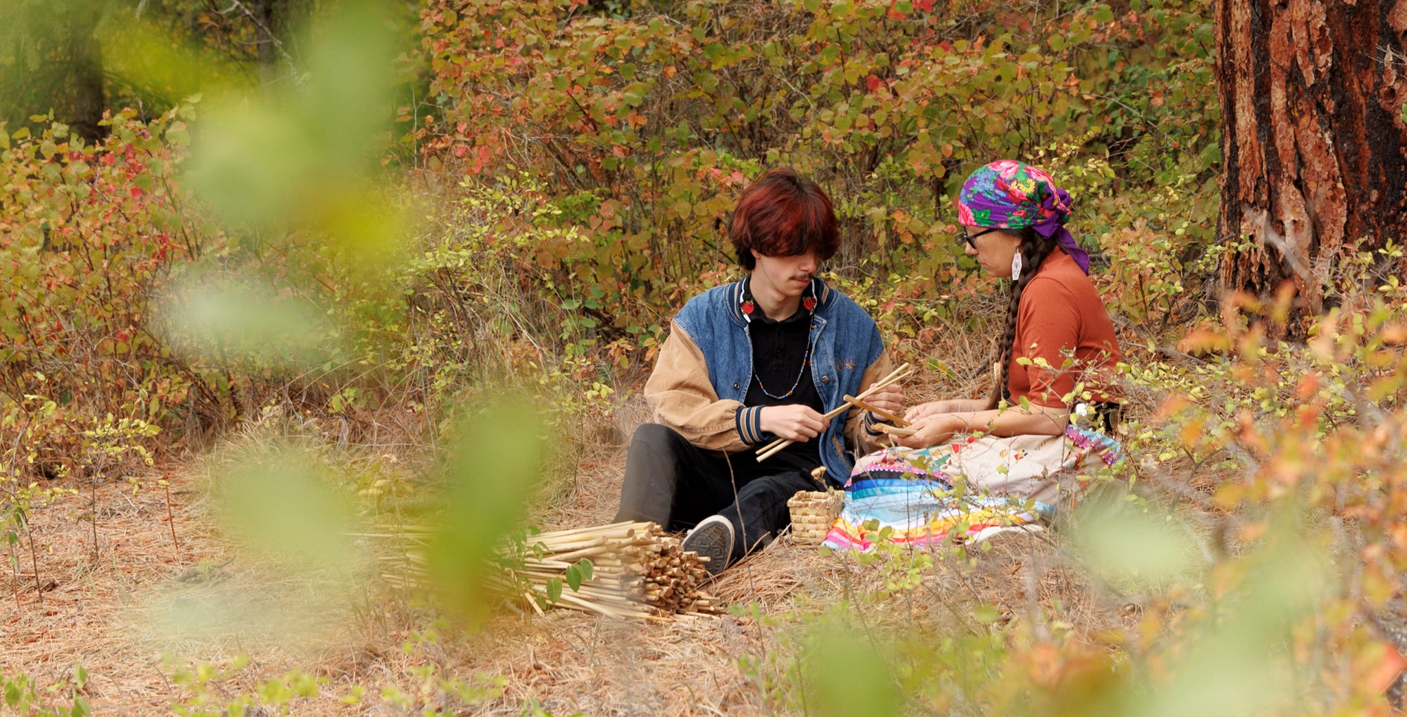 A photo of Neomia Battin and his auntie Alisha Athos sitting together on the forest floor, weaving a tule basket. A bundle of cut tule stalks and a small woven basket rest beside them.