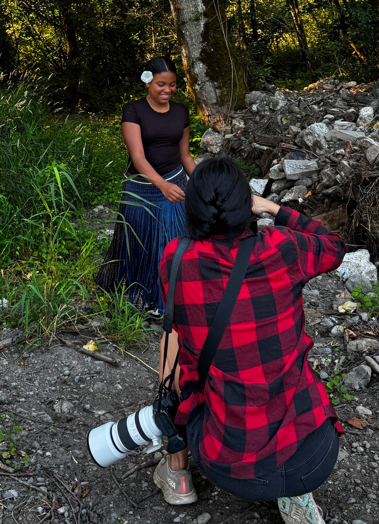 A behind-the-scenes photo from a photo shoot in a wooded area. Photographer Tailyr Irvine crouches on a rocky path, camera raised, as Shaliya Hawkins smiles at the edge of the trees wearing a white flower in her hair and a long woven skirt.