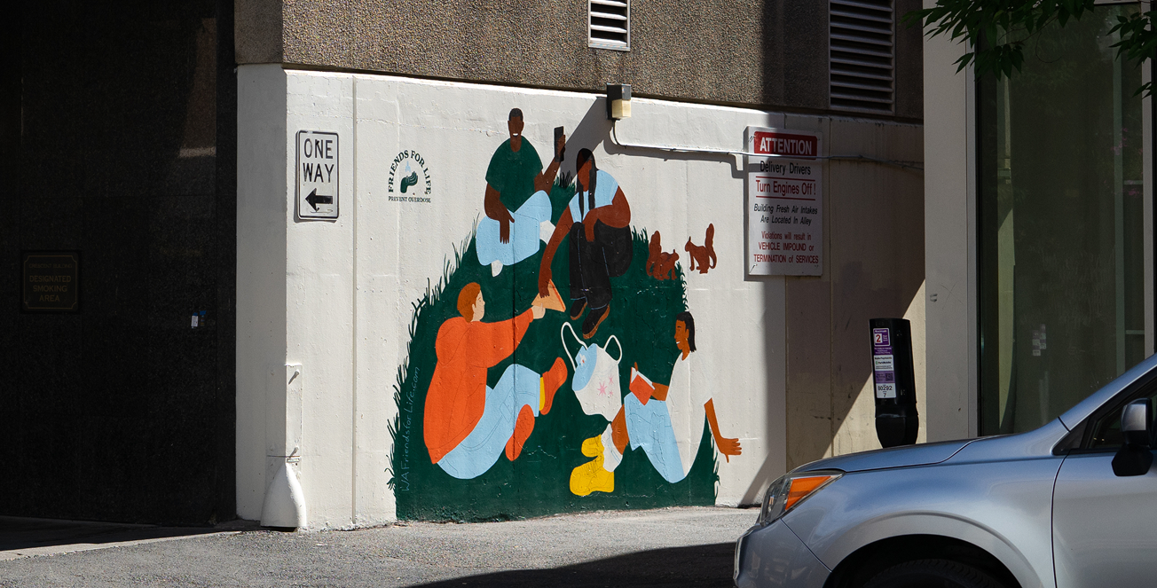 Colorful mural on a white exterior wall depicting a diverse group gathered around tote bag with naloxone in it, with a parked car partially blocking the lower-right corner.
