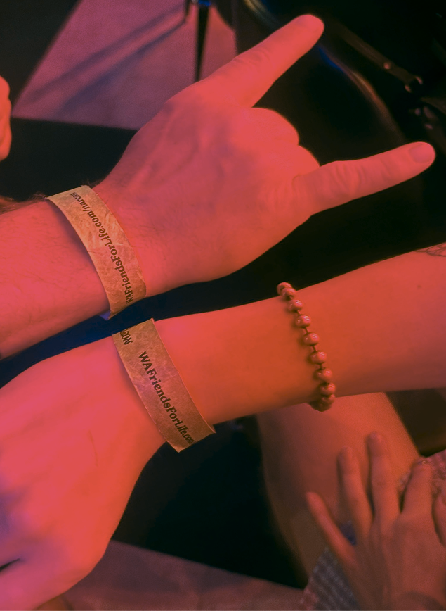Close-up of two forearms under red lighting wearing Friends for Life event wristbands, with one hand making a ‘rock on’ gesture.
