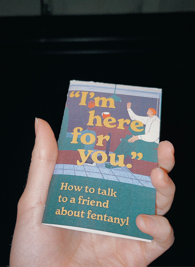 A hand holds a booklet titled “I’m here for you: How to talk to a friend about fentanyl” against a dark background.