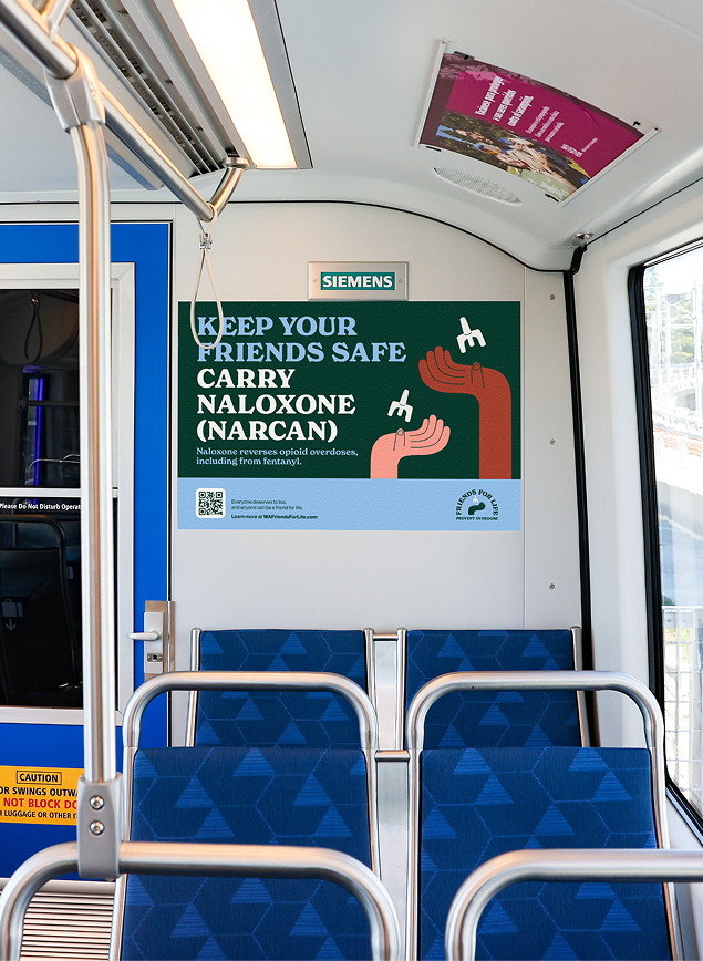 Naloxone safety poster above blue train seats reading, “Keep your friends safe. Carry naloxone (Narcan),” alongside illustrated hands. 