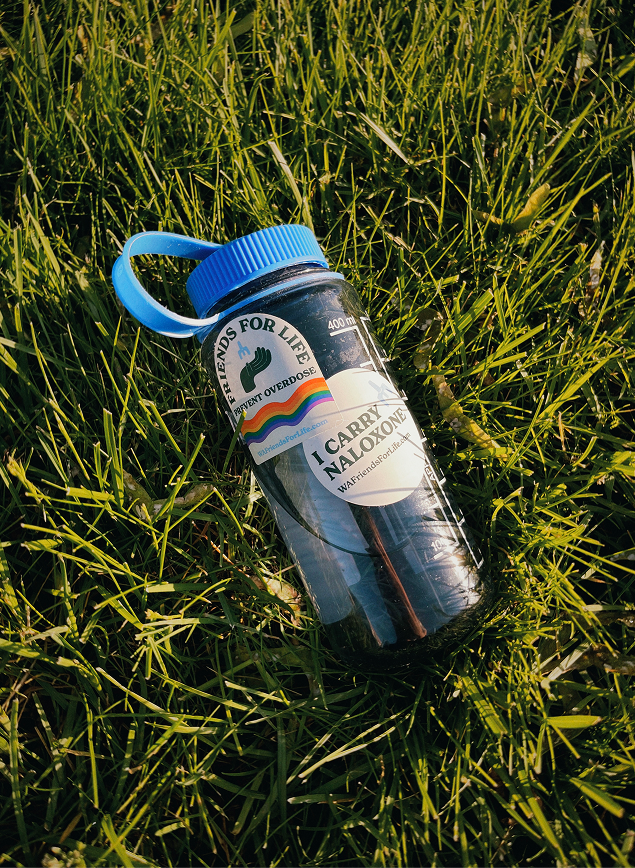 Clear water bottle with a blue cap lying in sunlit grass, decorated with “Friends for Life” and “I Carry Naloxone” stickers. 