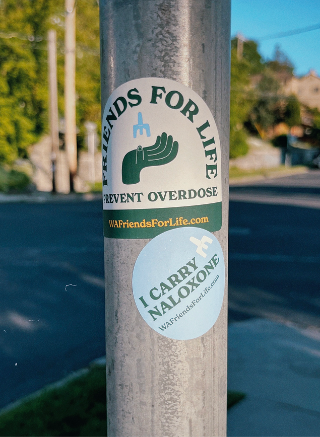 Two green-and-white overdose-prevention stickers on a metal street pole reading “Friends for Life” and “I Carry Naloxone,” with a residential street blurred behind them. 