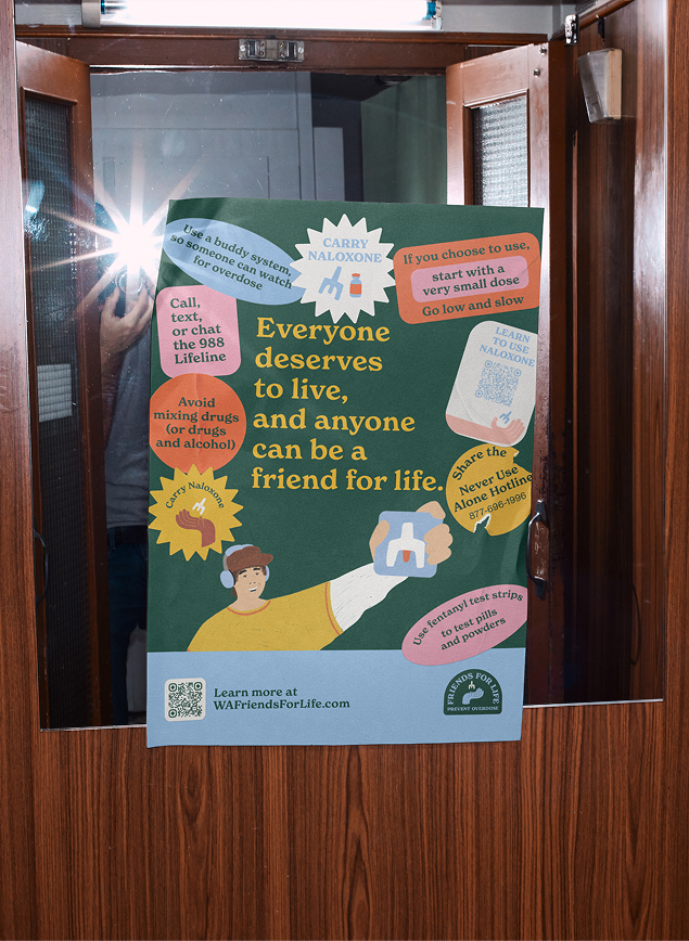 Overdose-prevention poster taped to a mirror beside a camera-flash reflection. Its central message reads, “Everyone deserves to live, and anyone can be a friend for life.” 
